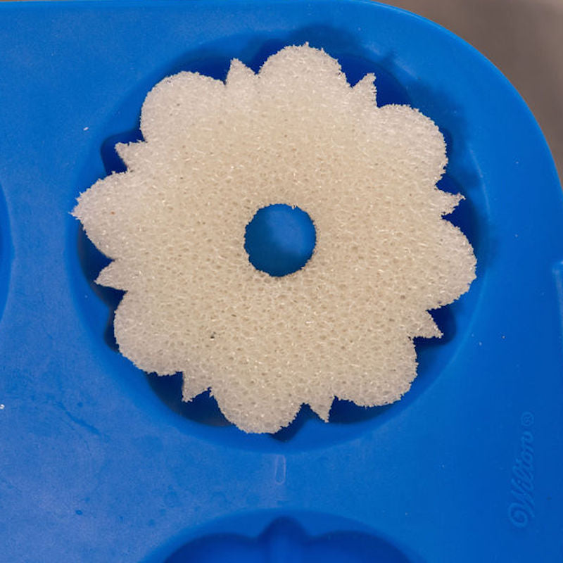 A close-up of a white, flower-shaped cleaning sponge with a central hole, resting inside a Wilton Bundt® pan