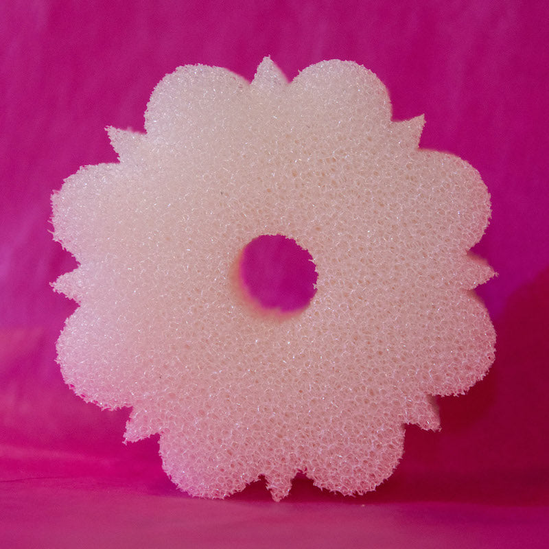 A flower-shaped cleaning sponge with a central hole for cleaning donut and Bundt pans set against a vibrant pink background.