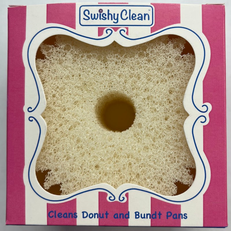 A white cleaning sponge with a central hole, displayed inside its square-shaped pink and white striped retail box with a decorative cutout and  labeled 'Swishy Clean' and 'Cleans Donut and Bundt Pans'