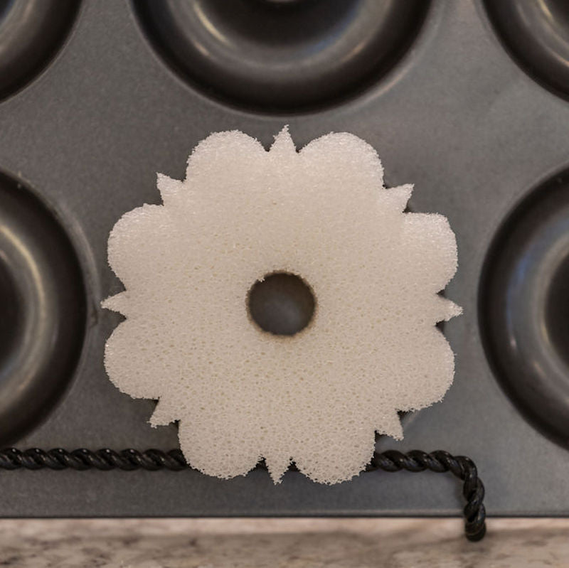 A white, flower-shaped cleaning sponge with a central hole, resting on a Wilton donut pan.