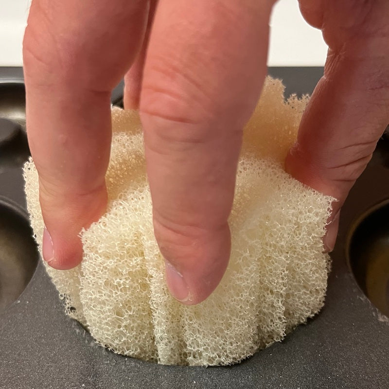 A close-up of a hand holding and pressing down on a white, porous cleaning sponge-placed over a donut pan.